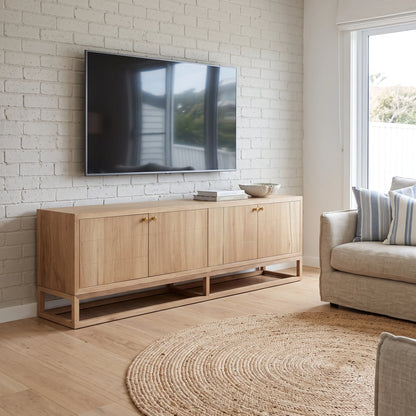 A light natural wood TV stand cabinet with four doors and small brass knobs, styled in a modern living room against a white brick wall with a mounted flat-screen TV, a beige sofa, and a round jute rug.