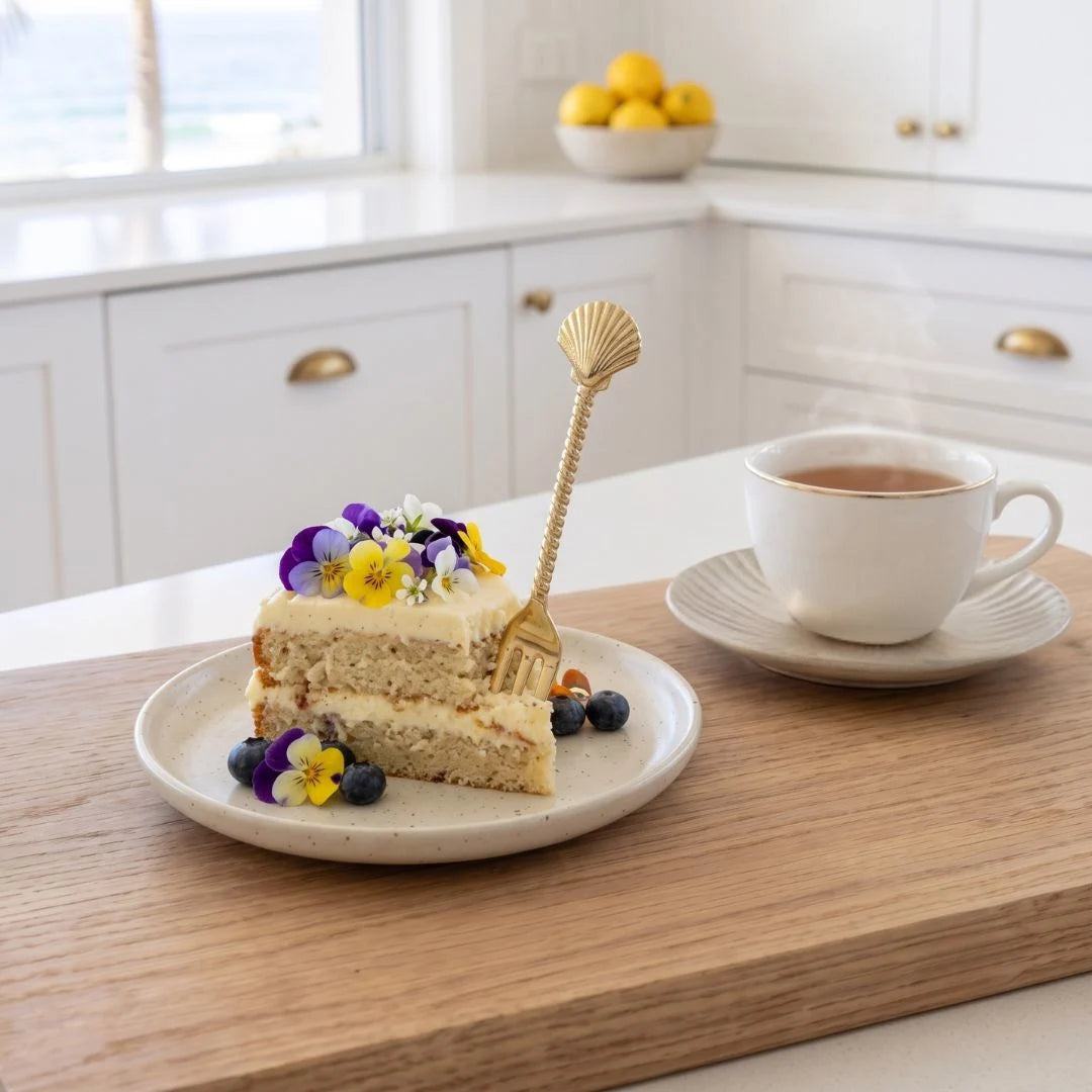A slice of layered cake on a small plate with edible flowers and blueberries, set on a wooden board beside a white cup of tea in a bright kitchen. A decorative gold shell-shaped fork rests on the cake.