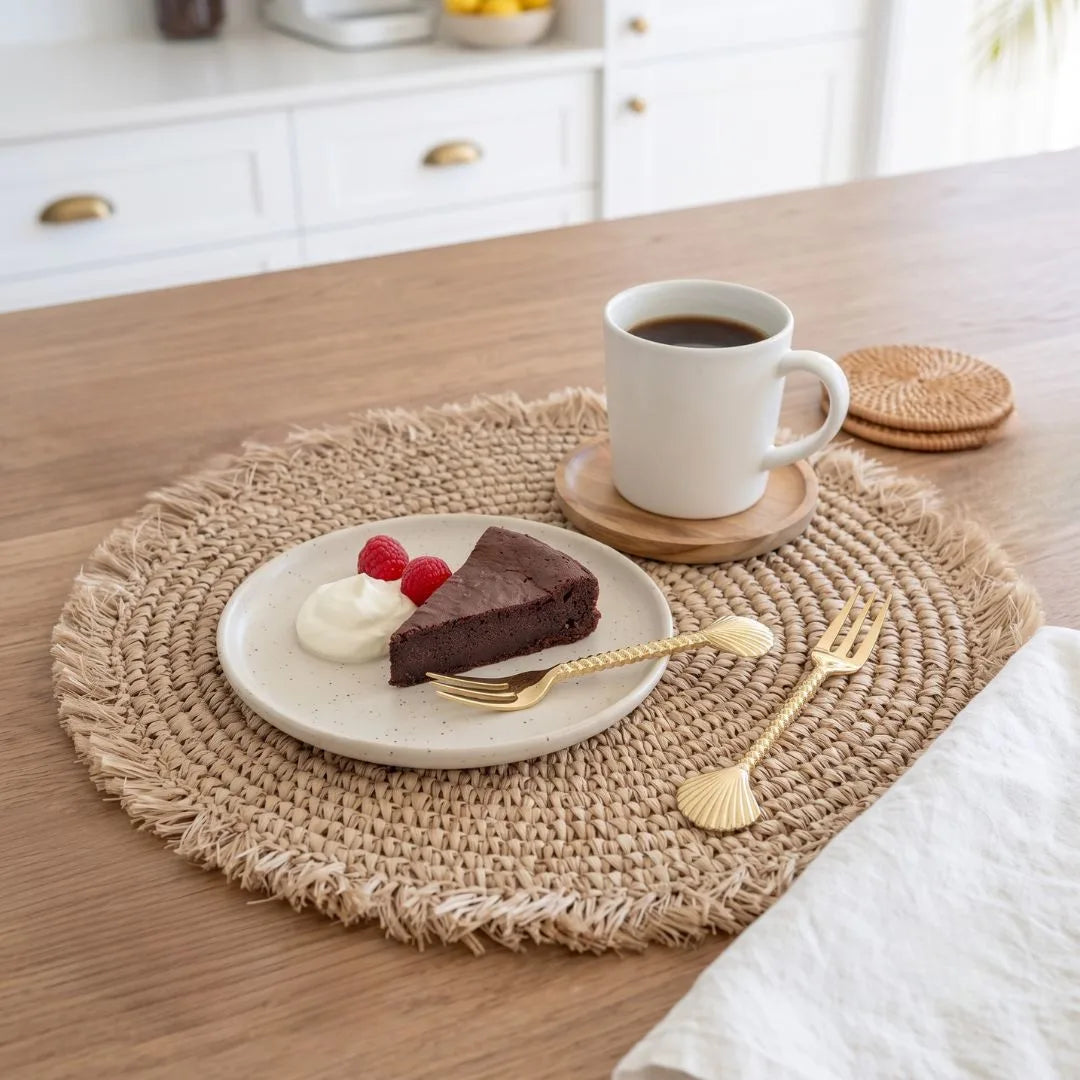 A round woven beige placemat on a wooden table styled with a plate of chocolate cake and whipped cream, a cup of coffee on a coaster, gold-toned shell-shaped forks, and a white napkin in a bright kitchen setting.