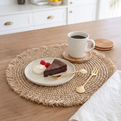 A round woven beige placemat on a wooden table styled with a plate of chocolate cake and whipped cream, a cup of coffee on a coaster, gold-toned shell-shaped forks, and a white napkin in a bright kitchen setting.