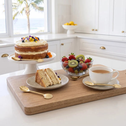 A styled breakfast scene in a bright white kitchen featuring a wooden serving board with a cup of tea, a slice of cake on a plate, a fruit bowl, and a cake on a pedestal stand, all arranged with gold-toned spoons.