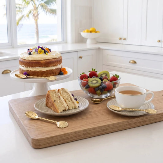 A styled breakfast scene in a bright white kitchen featuring a wooden serving board with a cup of tea, a slice of cake on a plate, a fruit bowl, and a cake on a pedestal stand, all arranged with gold-toned spoons.