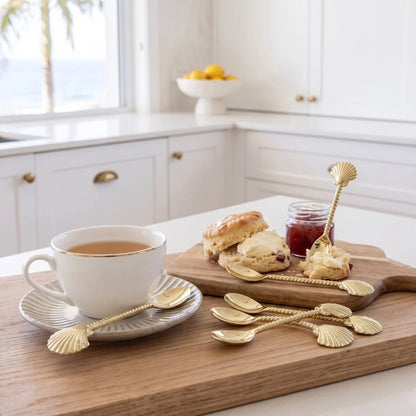 A styled kitchen scene showing a white teacup and saucer with a gold leaf-shaped spoon, a wooden serving board with scones, jam, and additional gold leaf spoons, and a blurred kitchen background with a window and ocean view.