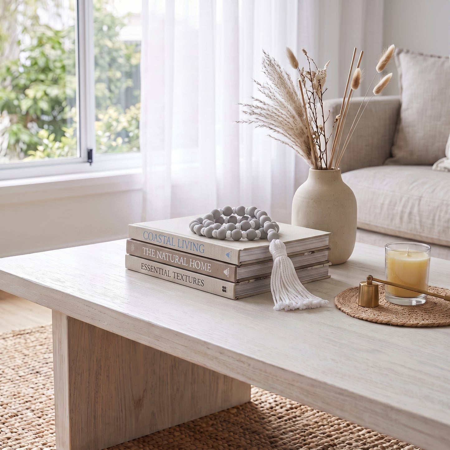 A styled living room scene featuring a light wood coffee table with stacked books, a beaded ornament, a white tassel, a beige ceramic vase with dried grasses, and a candle on a woven coaster. A window, sheer curtains, and a neutral sofa are visible in the background.