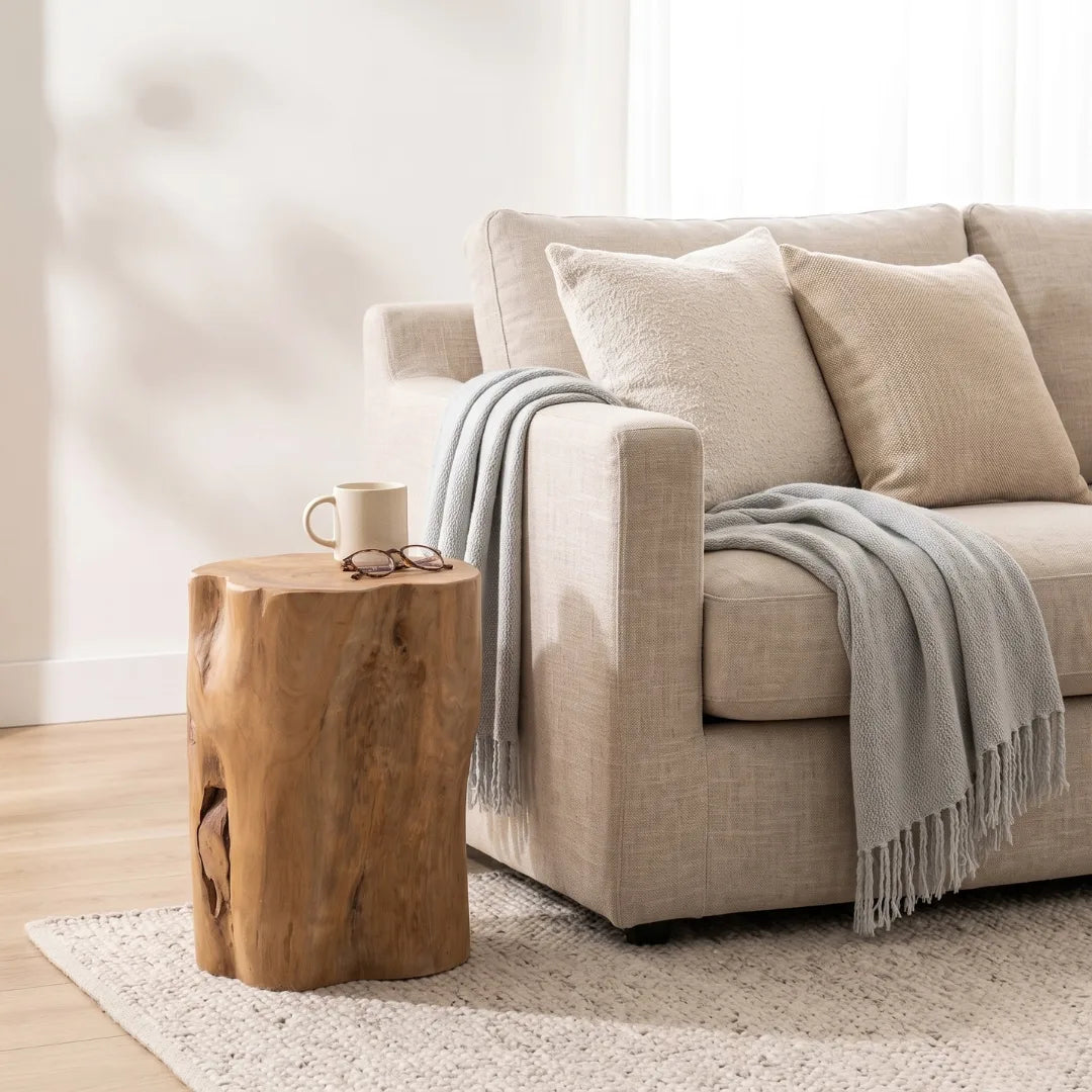 A light beige upholstered sofa in a bright living room, styled with cream pillows and a gray throw blanket. A rustic wooden side table sits beside the sofa holding a mug and eyeglasses.
