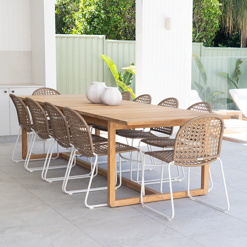 A rectangular wooden dining table with woven rattan chairs, set on a light gray tiled patio under a covered outdoor area with a glass fence and lounge chair in the background.