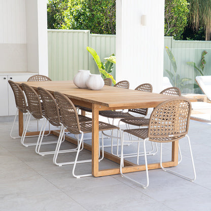 A rectangular wooden dining table with woven rattan chairs, set on a light gray tiled patio under a covered outdoor area with a glass fence and lounge chair in the background.