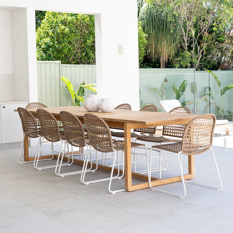 A modern outdoor dining set featuring a rectangular light oak wood table and eight woven rattan chairs with white metal legs, set on a light gray tiled patio under a covered outdoor area.