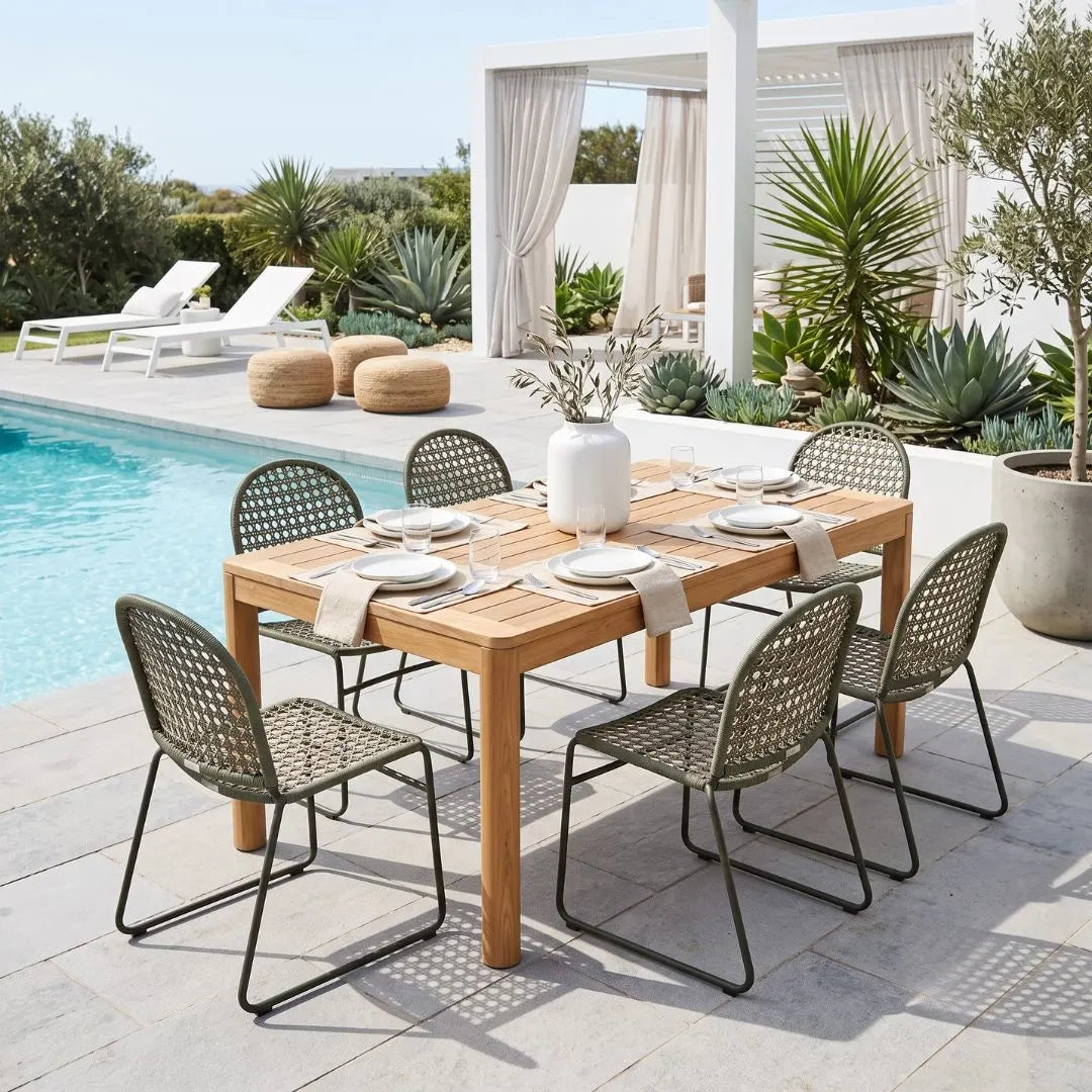 A rectangular wooden dining table set with twelve woven metal chairs, place settings, and a large greenery centerpiece, positioned outdoors on a patio next to a swimming pool and modern house.