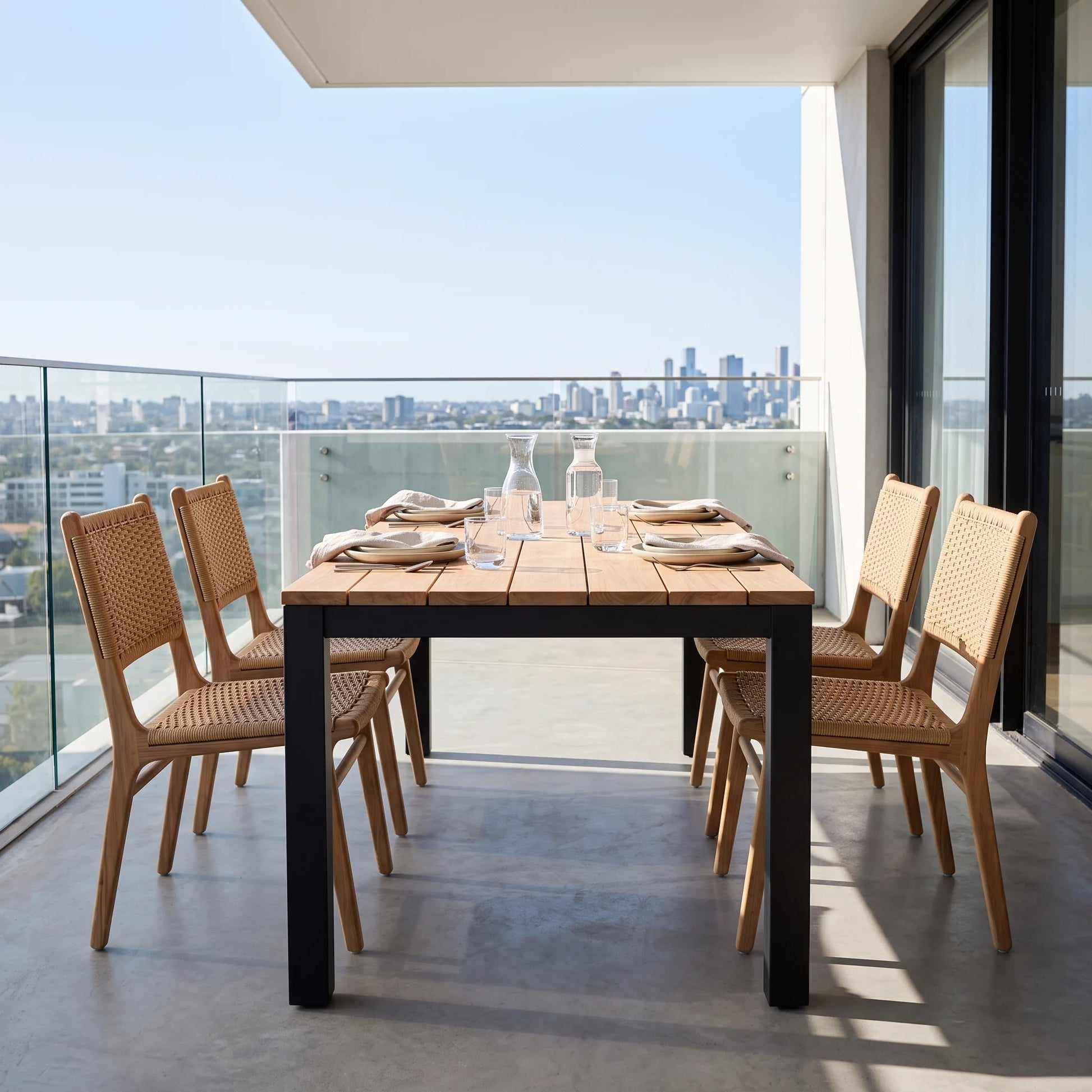 Outdoor dining set on a balcony featuring a rectangular wooden table with black legs and four matching woven-back chairs, set against a city skyline background.