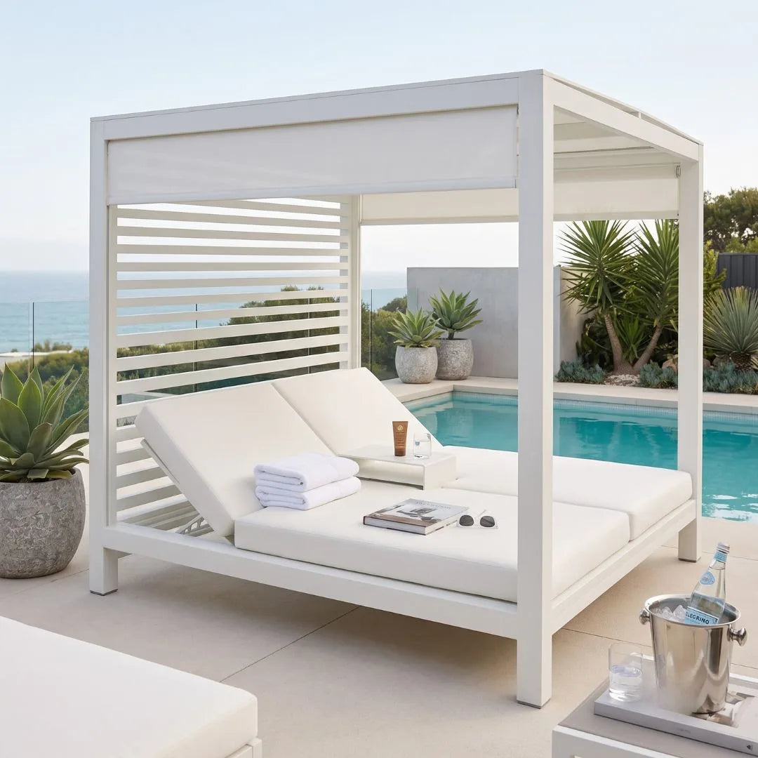 A white outdoor cabana-style lounge chair with thick cream cushions, set beside a swimming pool and ocean view. A small table in front holds an ice bucket with a bottle and glasses, and the chair is styled with folded white towels, a small cup, and a book.