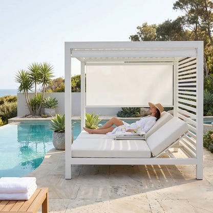 A woman relaxes on a white outdoor cabana daybed with a poolside setting, featuring a white frame, white cushions, a retractable white shade, and a stone patio background.