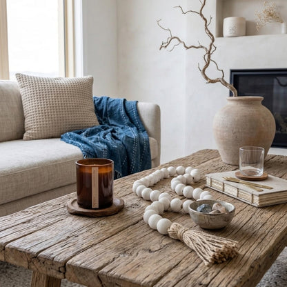 A rustic wooden coffee table in a cozy living room setting, styled with a white beaded tassel necklace, a brown amber glass candle, a small bowl, and stacked books. In the background, a beige sofa with a textured pillow and a blue throw blanket is visible next to a fireplace.
