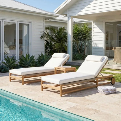 Two wooden outdoor chaise lounge chairs with white cushions are positioned on a light stone patio beside a swimming pool, with a white house and green plants in the background.
