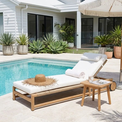 A modern outdoor chaise lounge with a light wood frame and white cushion sits beside a swimming pool on a tiled patio. A rolled white towel rests on the footrest, a woven straw hat and small side table with a glass of water are placed nearby, and a white house with large windows and potted plants is in the background.