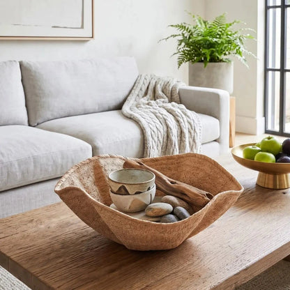 A large, organic-shaped woven wood bowl sits on a rustic wooden coffee table in a bright, modern living room. The bowl contains a small ceramic cup, driftwood, and smooth stones.