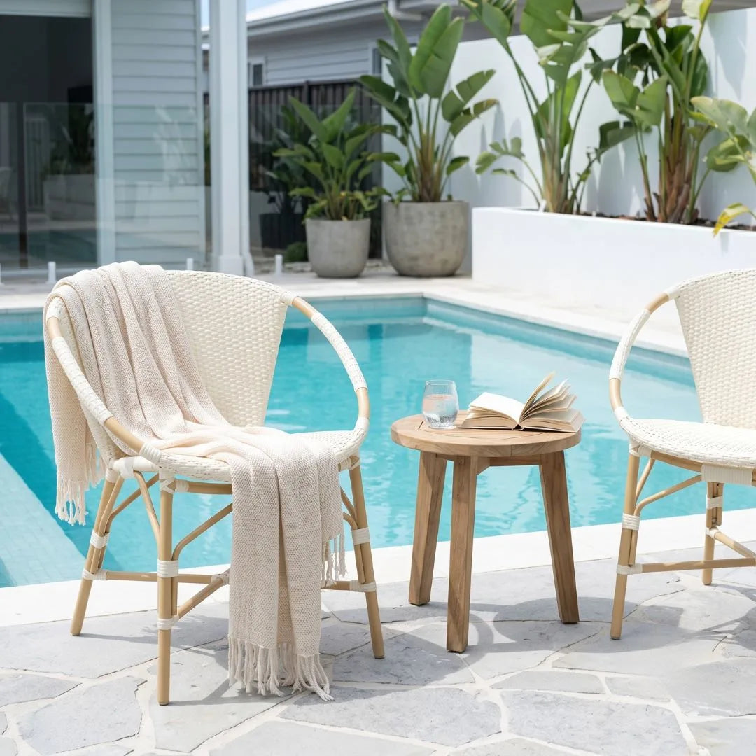 Two woven outdoor patio chairs with a small round wooden side table between them, set beside a swimming pool on a stone patio. A cream throw blanket is draped over the left chair, and an open book and glass are on the table.