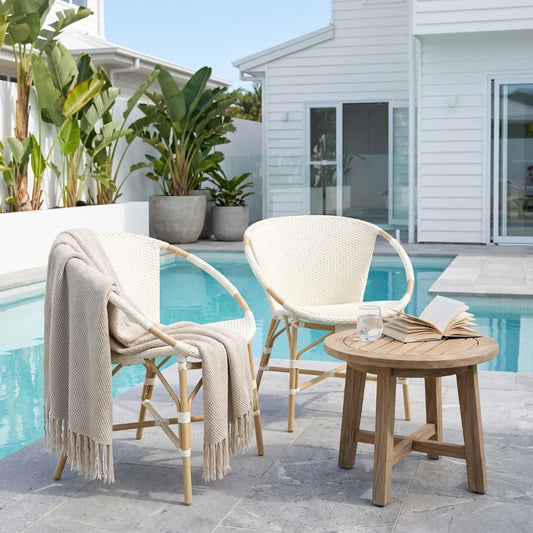 Outdoor patio furniture set with two woven armchairs, a round wooden side table, and a beige throw blanket draped over one chair, set beside a swimming pool in front of a white house.