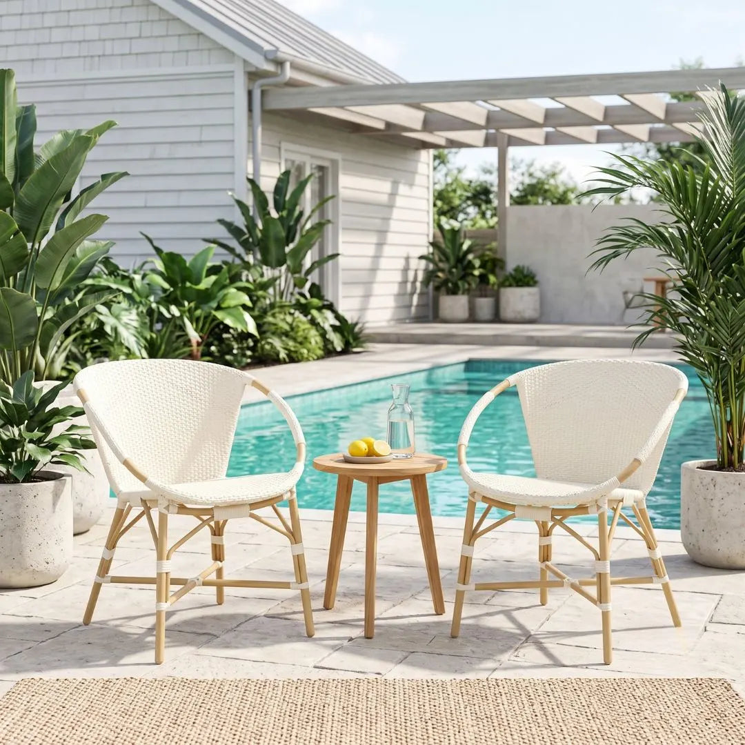 Two cream woven outdoor dining chairs with light wood frames and a small round wooden side table between them, set on a patio poolside with a house, plants, and pergola in the background.