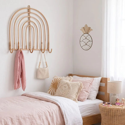A light natural wood wall-mounted arched rack with five hooks is shown in a styled bedroom setting, holding a pink towel and a cream woven handbag. The room features a wooden bed frame, pink and cream bedding, a woven pineapple wall decor piece, and a wicker side table with a lamp.
