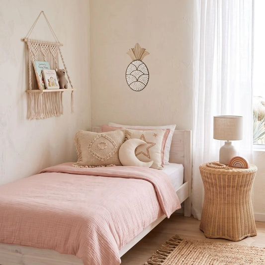 A styled children's bedroom featuring a single bed with a dusty pink quilt, decorative pillows, a woven macrame wall hanging, a wicker side table with a lamp, and a pineapple wall decor piece.