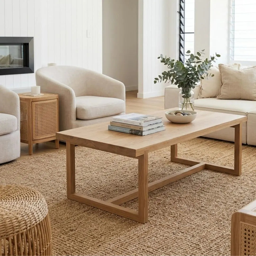 A light natural wood rectangular coffee table with a geometric frame base, styled in a bright, modern living room with a woven jute rug, cream armchair, and side table.