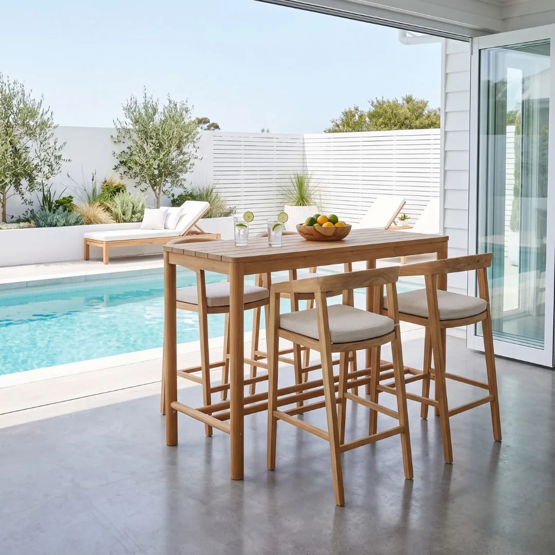A wooden outdoor bar-height dining set with a rectangular table and four matching stools with light beige cushions, set on a concrete patio beside a swimming pool and white slatted fence.