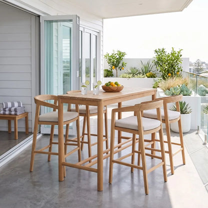 A wooden outdoor bar-height dining set with a rectangular table and four matching stools with light beige upholstered seats, placed on a balcony with plants and sliding glass doors in the background.