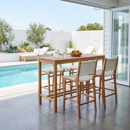 A wooden outdoor bar-height dining table with four woven-back bar stools, set on a concrete patio next to a swimming pool with a white slatted fence and lounge chairs in the background.