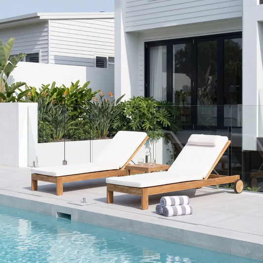 Two wooden outdoor chaise lounge chairs with white cushions are positioned beside a swimming pool on a light stone patio, with a modern white house and green plants in the background.