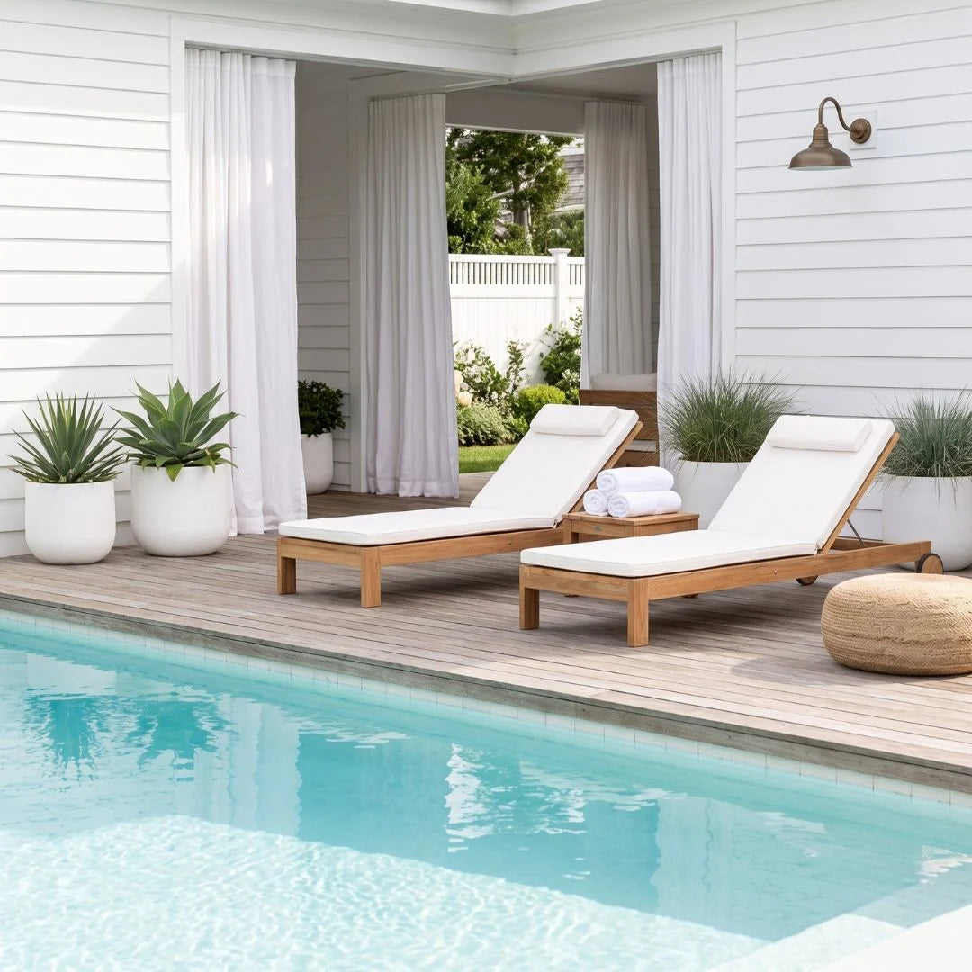 Two wooden outdoor chaise lounge chairs with white cushions sit on a wooden deck beside a swimming pool, with white house siding, potted plants, and a covered patio in the background.