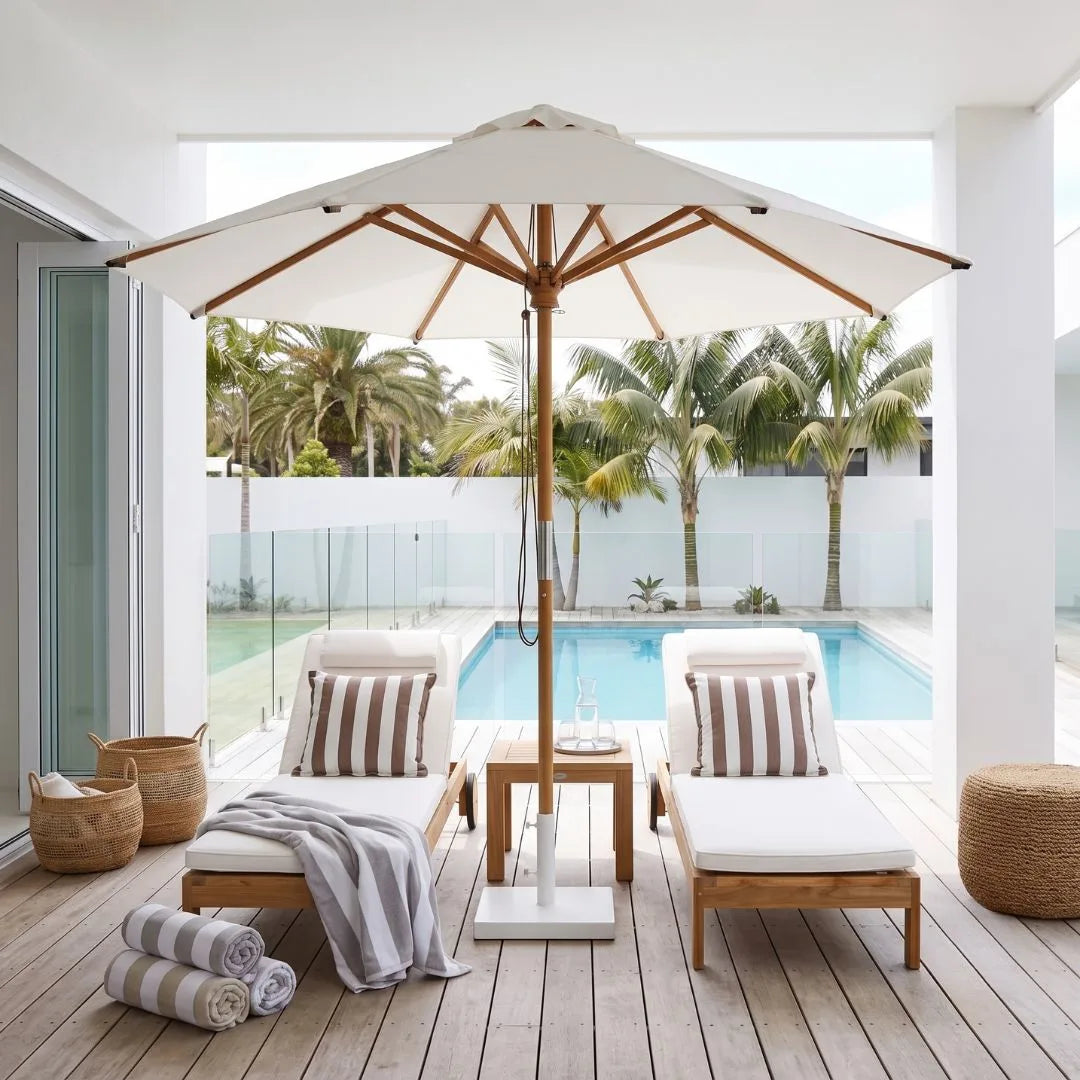 A luxury outdoor patio setup featuring a large cream-colored patio umbrella with a natural wood frame, two wooden lounge chairs with striped cushions, a small wooden side table, and a view of a swimming pool and palm trees in the background.