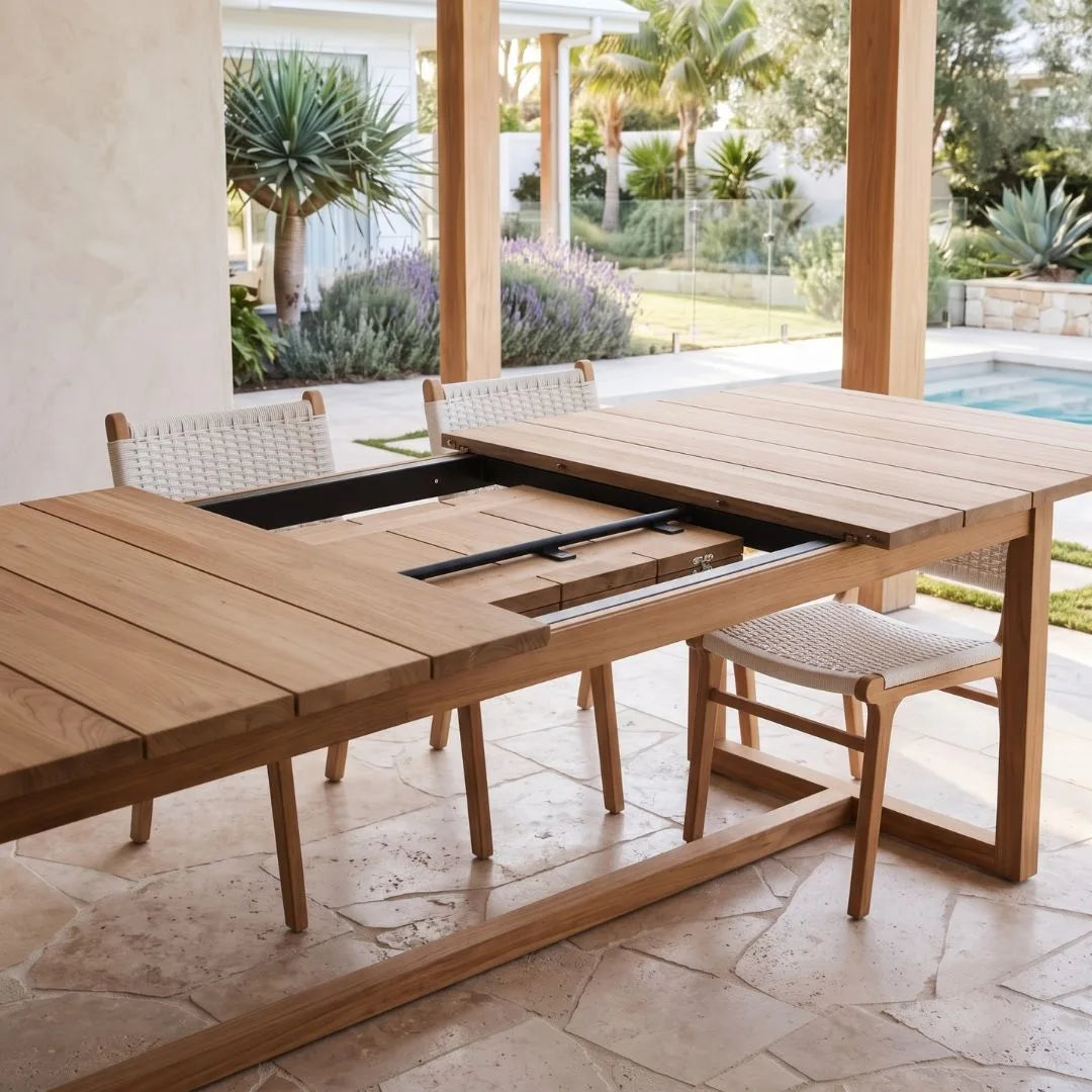 A wooden outdoor dining table with an inset dining area and woven chairs, shown on a covered patio with a pool and landscaped garden in the background.