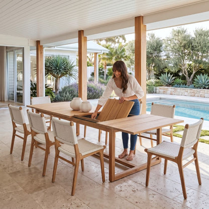 A woman sets a wooden dining table on a covered patio next to a swimming pool, with six matching wooden chairs featuring woven seats, in a bright, modern outdoor setting.