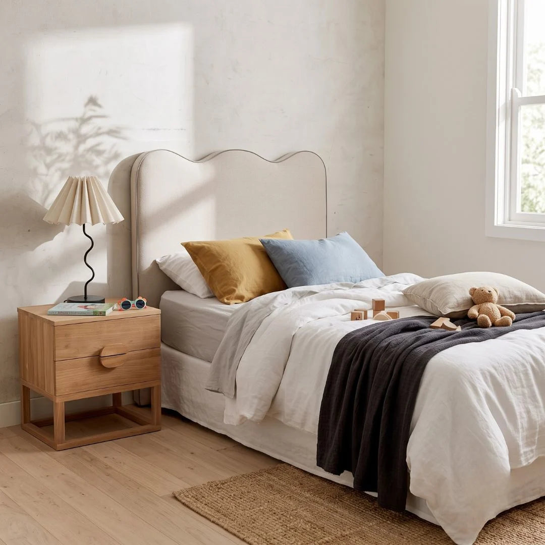 A styled bedroom scene featuring a bed with white linens, mustard yellow and light blue pillows, a dark charcoal throw blanket, a wooden nightstand with a pleated lamp, and a textured wall with soft shadows.