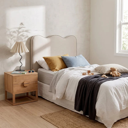A styled bedroom scene featuring a bed with white linens, mustard yellow and light blue pillows, a dark charcoal throw blanket, a wooden nightstand with a pleated lamp, and a textured wall with soft shadows.