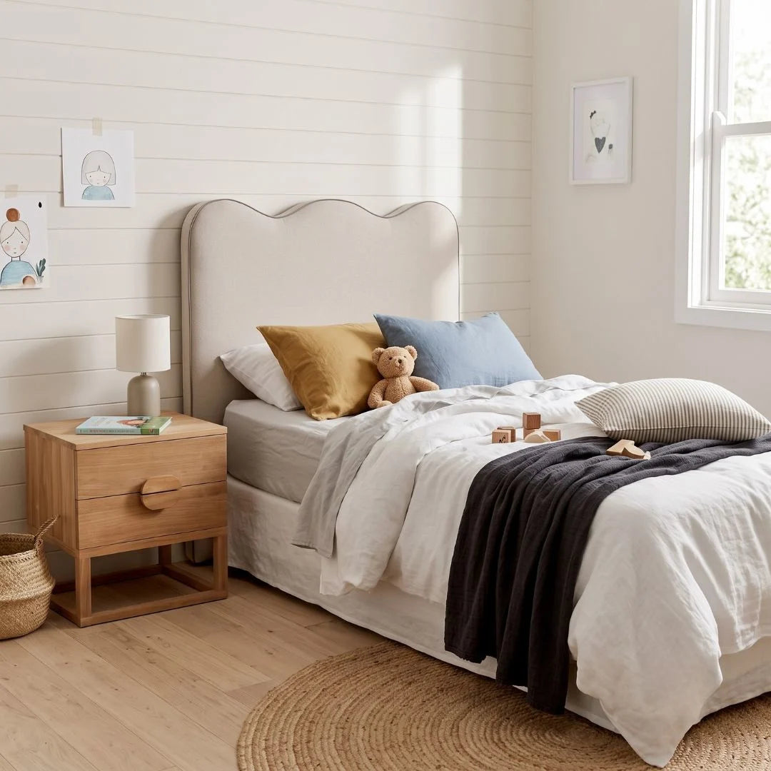 A styled bedroom scene showing a bed with a beige upholstered headboard, white bedding, mustard yellow and blue pillows, a teddy bear, a wooden nightstand, and a window with natural light.