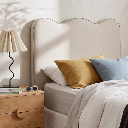 A beige upholstered headboard with a scalloped top edge in a styled bedroom setting, shown with white bedding, a mustard yellow pillow, and a light blue pillow on a wooden nightstand with a pleated lamp.