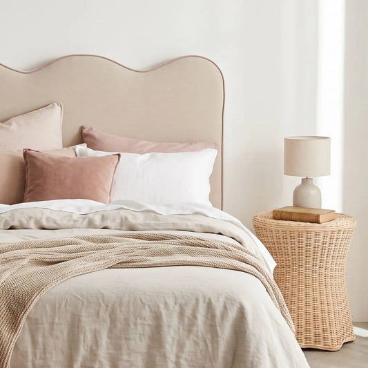 A softly styled bed with a wavy upholstered headboard in a warm beige tone, dressed in neutral linens and pillows including blush, white, and taupe. A woven side table beside the bed holds a small lamp and book in a bright, minimal bedroom setting.