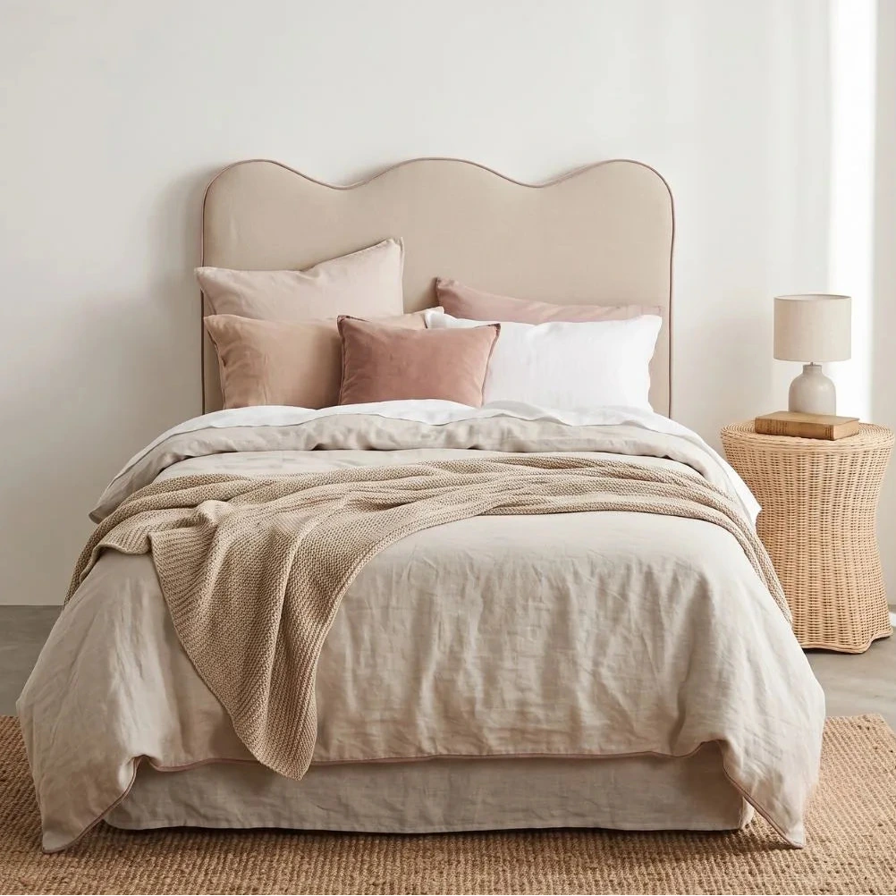 A neatly made bed with a wavy upholstered headboard, dressed in cream and blush bedding with matching pillows, set in a softly lit bedroom with a woven side table and lamp.