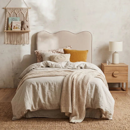 A neatly made bed in a softly styled bedroom, featuring a light beige upholstered headboard, cream bedding, patterned pillows, a mustard yellow accent pillow, a textured cream throw blanket, a wooden nightstand with a white lamp, and a macrame wall hanging.