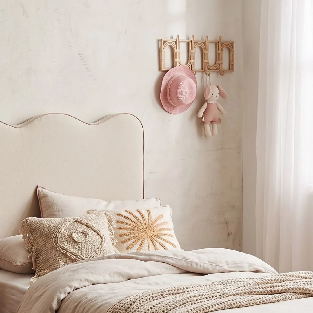 A styled bedroom corner with a cream upholstered bed, neutral bedding, decorative pillows, a pink hat, a small plush bunny, and a wall-mounted wooden rack on a light textured wall.