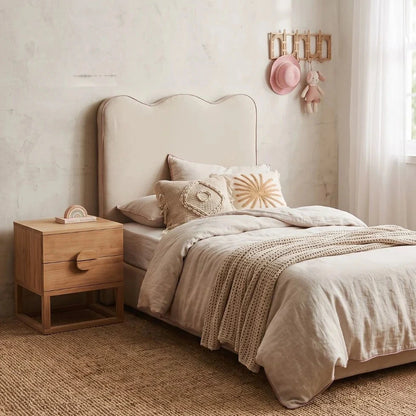 A styled bed in a softly lit bedroom featuring a cream upholstered headboard, neutral beige bedding, textured throw blankets, a small wooden nightstand, and a window with sheer curtains.