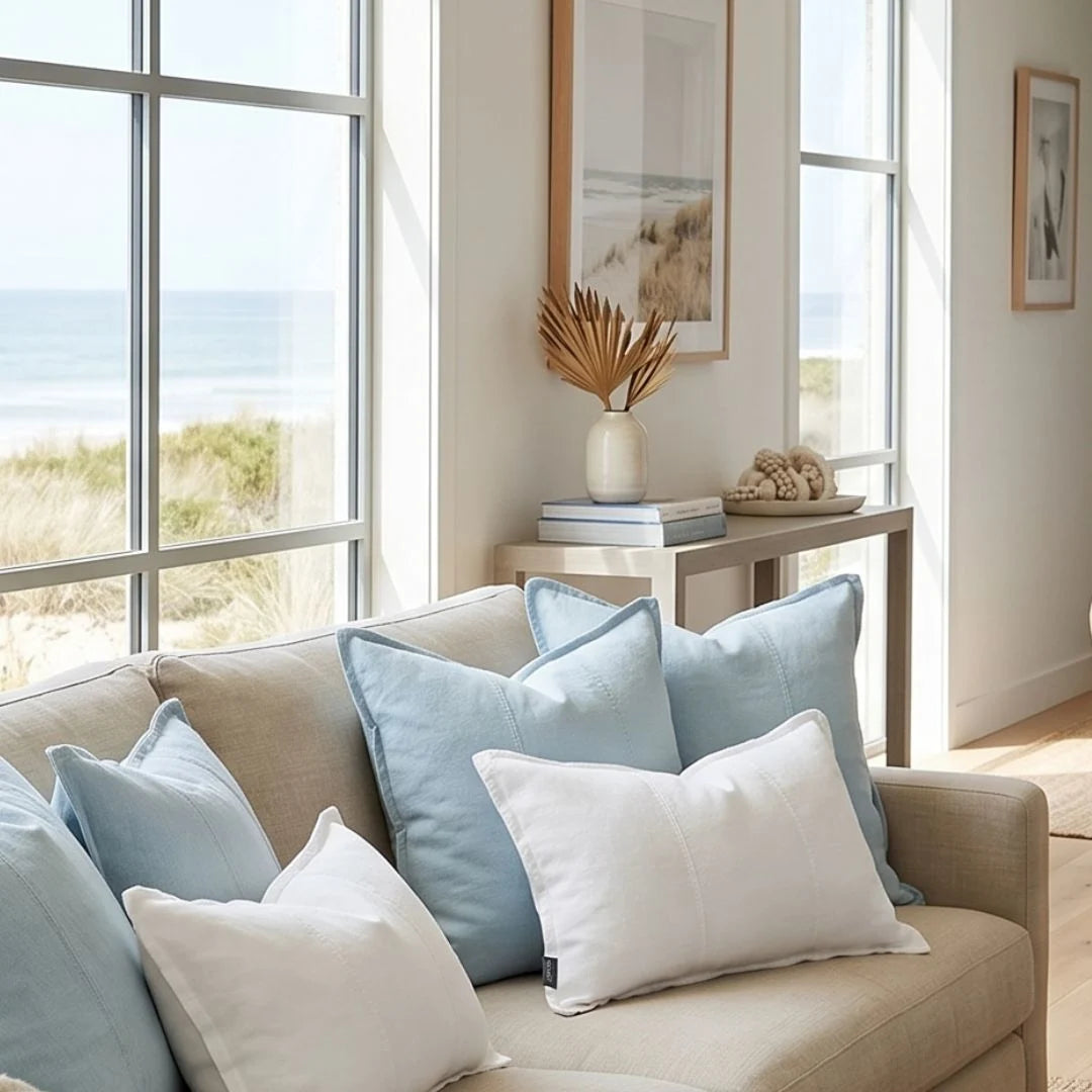 Beige sofa with light blue and white pillows in a room with large windows showing a beach view.