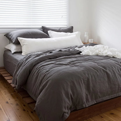 A neatly made bed with dark gray bedding and white pillows in a bright bedroom with wooden flooring and white blinds.