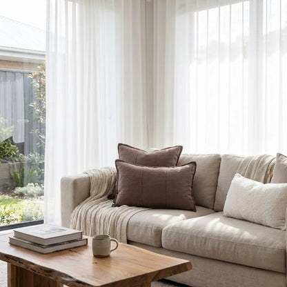 Beige sofa with brown and white cushions in a bright living room with large windows.