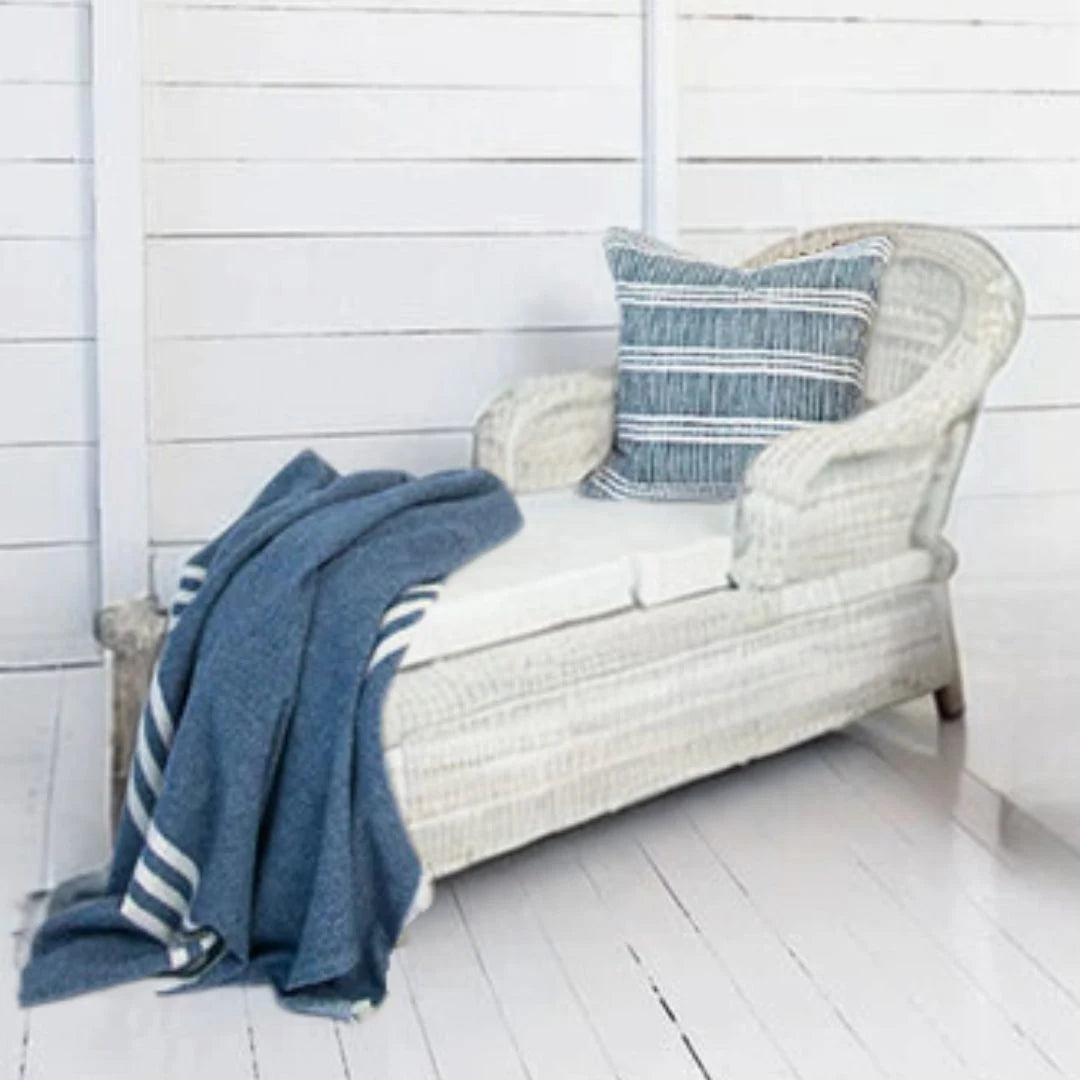 White wicker bench with blue striped pillow and blanket against a white wooden wall.