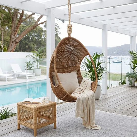 Hanging wicker chair with a blanket on a wooden deck by a poolside.
