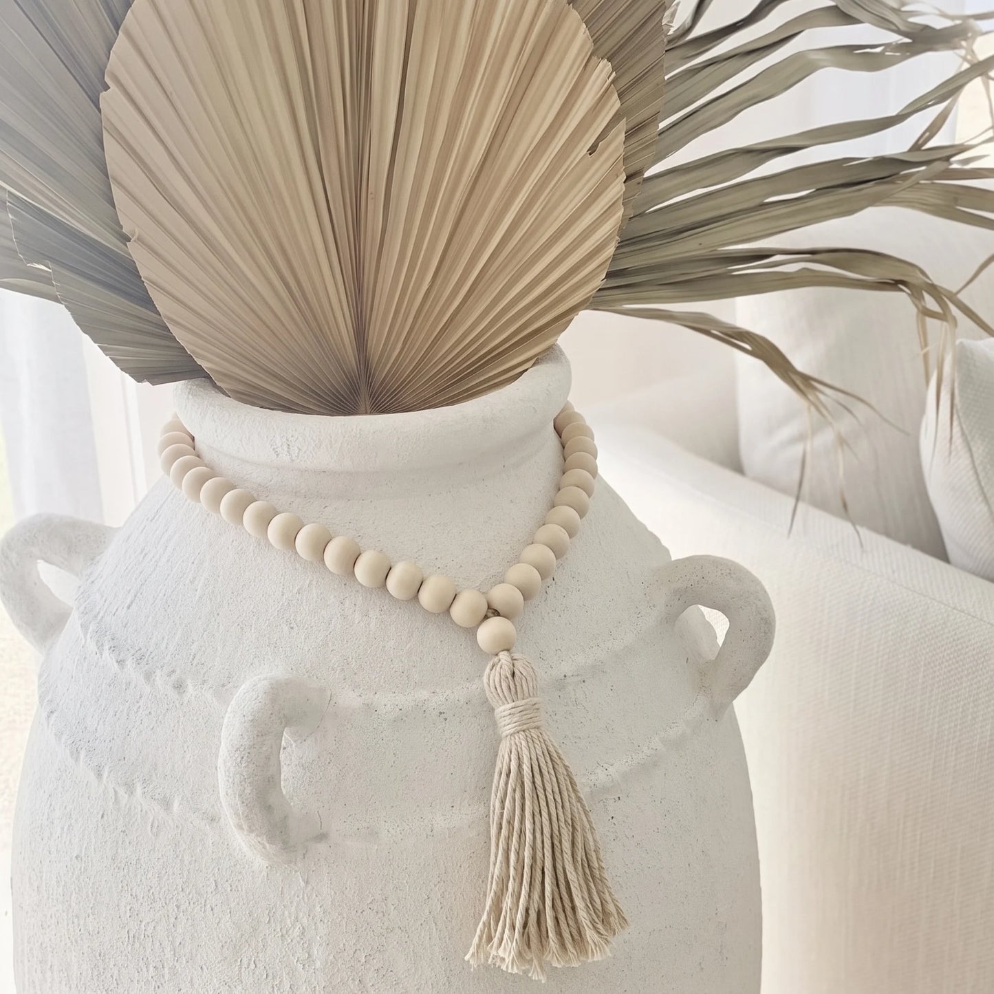 A natural wood bead necklace with a tassel is draped around a white ceramic vase, styled with dried palm leaves in a bright, neutral indoor setting.