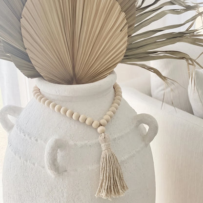 A natural wood bead necklace with a tassel is draped around a white ceramic vase, styled with dried palm leaves in a bright, neutral indoor setting.